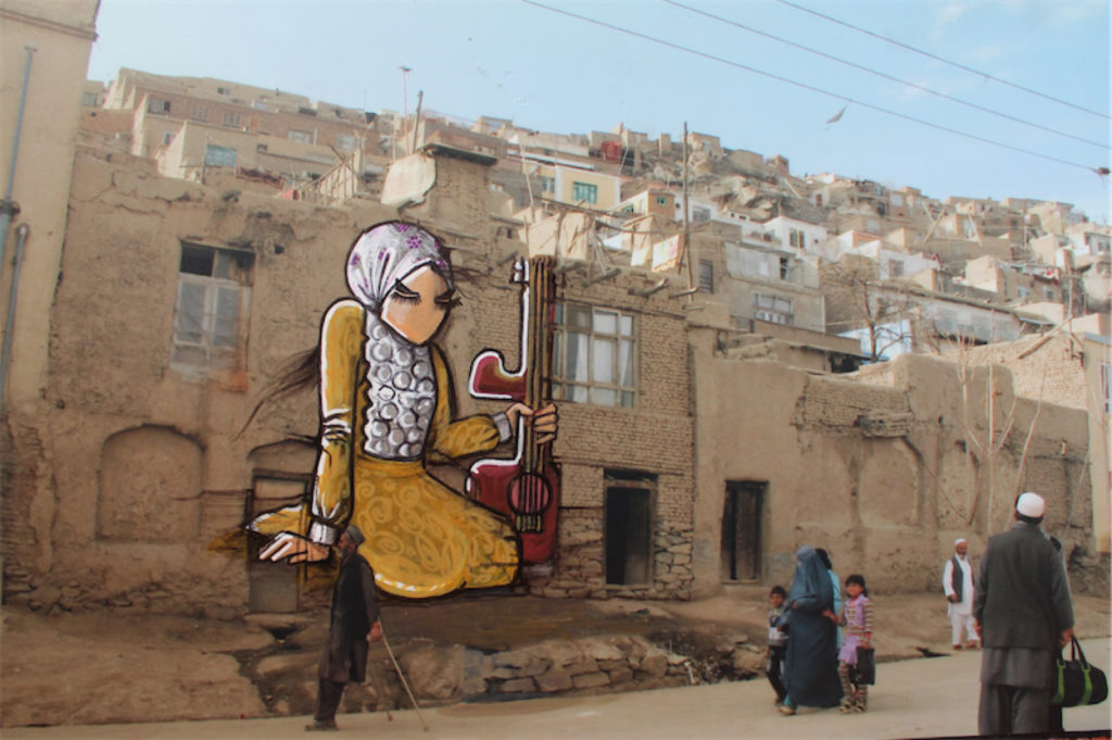 Graffiti of a woman in yellow, wearing a white headscarf and holding a guitar, on the walls of old houses in Afghanistan, with people walking by on the street.