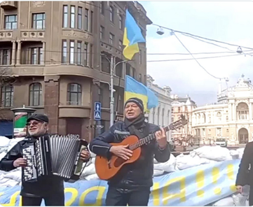 Photo of two Ukrainian men playing an accordion and guitar in front of sandbags in a city block. with Ukrainian flags in the background